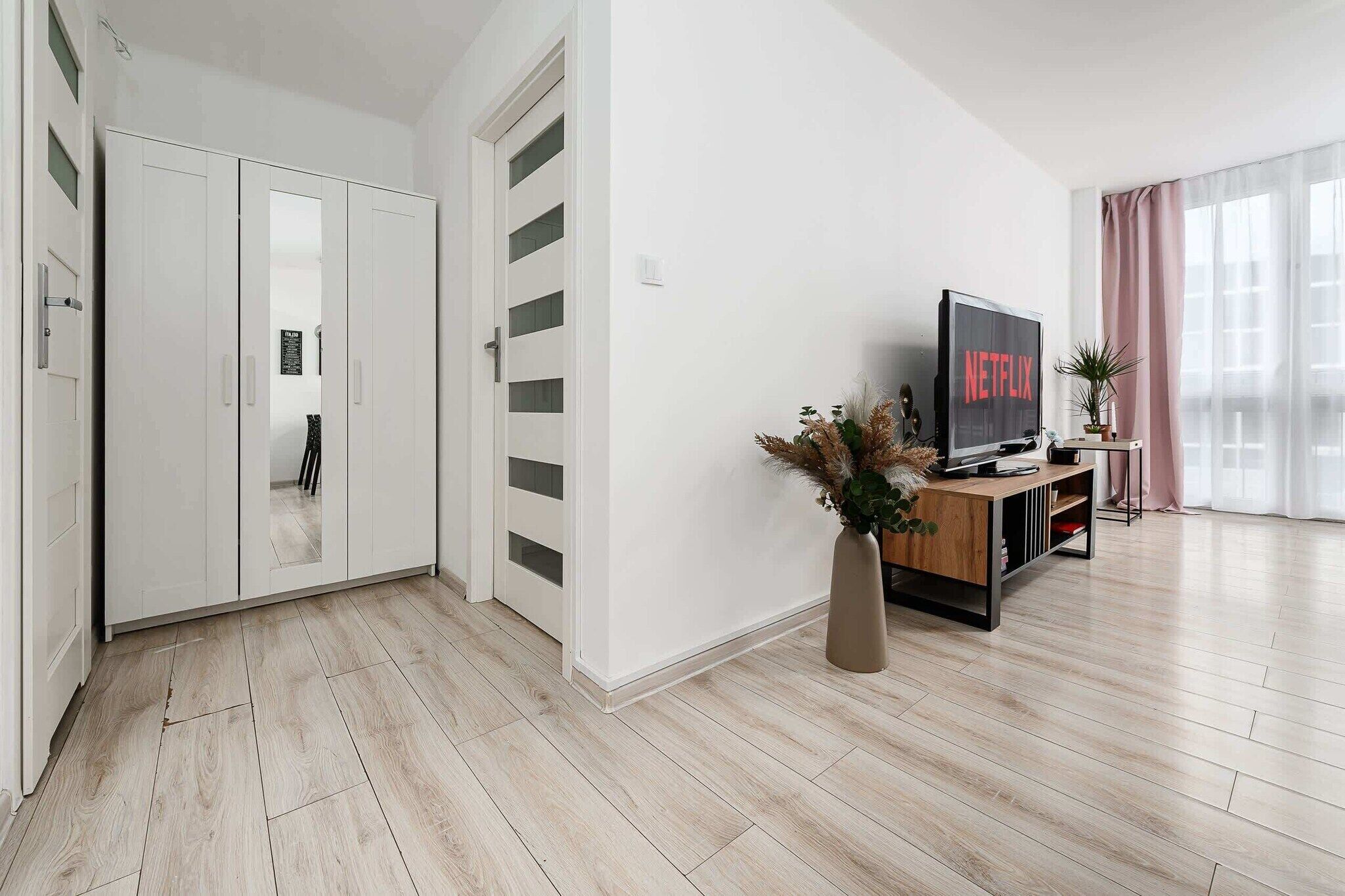 A modern living room with a wooden floor, white walls, a TV on a wooden stand, and a large window with curtains.