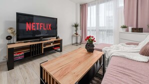 Un soggiorno moderno con una TV a schermo piatto che mostra 'NETFLIX', un tavolino in legno, un divano con una coperta bianca e una pianta in vaso.