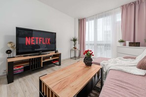 Ein modernes Wohnzimmer mit einem Flachbildfernseher, der 'NETFLIX' anzeigt, einem Holztisch, einer weißen Couch mit einer dekorativen Decke und einer Zimmerpflanze.