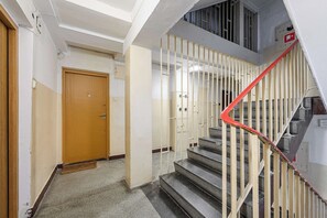 A staircase with a red handrail leading to an upper floor, a wooden door at the bottom, and a fire alarm on the wall. - Cozy Apartment for 6 near VIstula River (Warszawa 2)