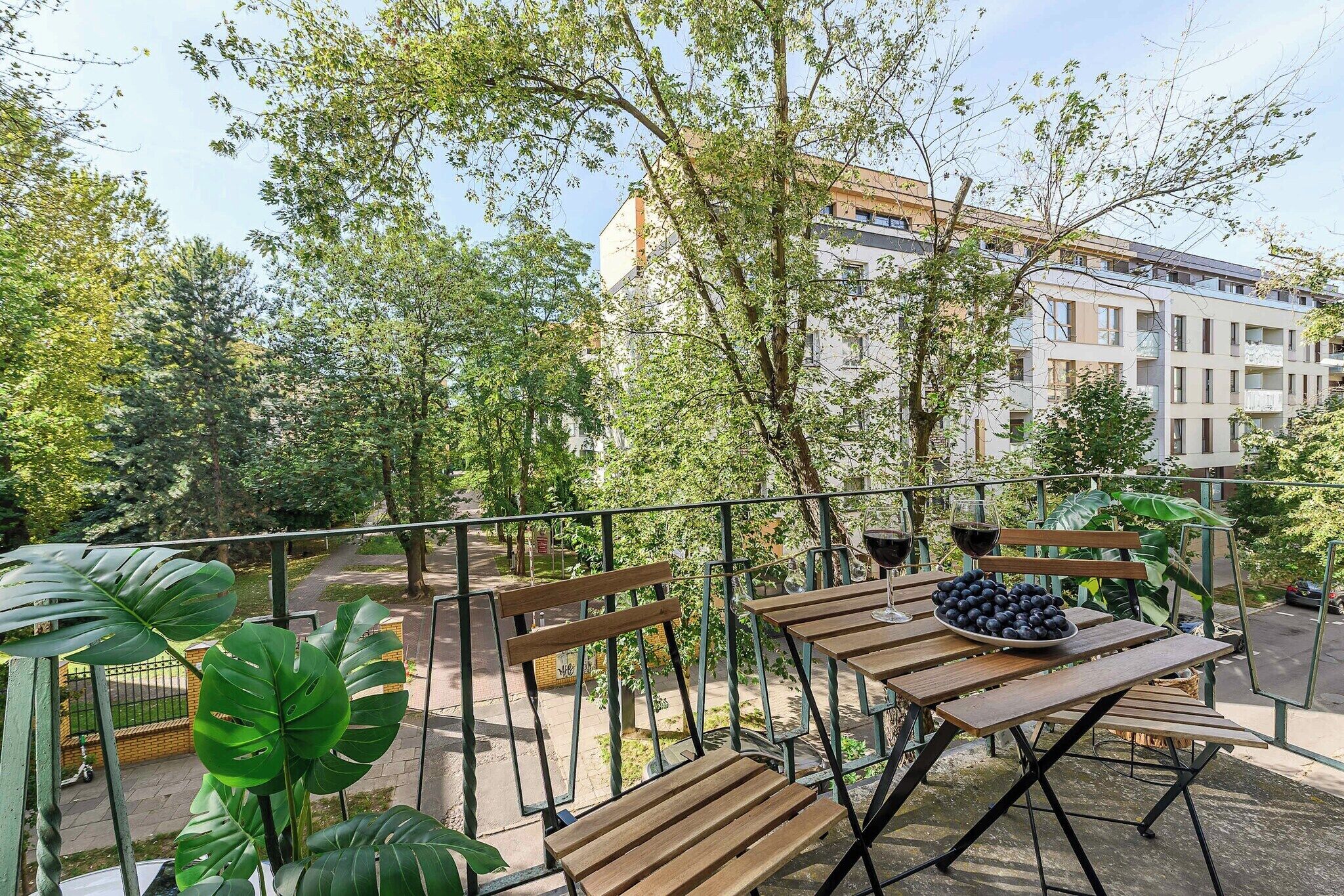 An outdoor seating area with wooden tables and benches, surrounded by greenery and a building in the background.