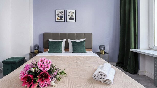 A hotel room with a bed, a nightstand, a bedside table, a window with curtains, and a wall with two framed pictures. - Lush Flat in Lodz Center (Łódź)