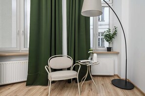Ein Zimmer mit einem grünen Vorhang, einem weißen Stuhl, einem kleinen Tisch mit einer Lampe und einer Topfpflanze.