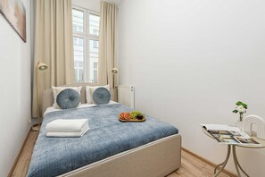 Ein ordentlich eingerichtetes Schlafzimmer mit Bett, Nachttischlampen, einem kleinen Tisch mit Pflanze und einem Fenster mit Vorhängen.