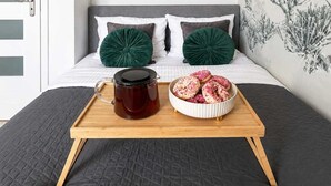A bed with a dark quilted bedspread, a wooden tray with a dark mug and a bowl of pastries, and two green pillows on a grey headboard. - Elegant Apartment near Vitsula River (Warszawa)