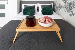 A bed with a dark quilted bedspread, a wooden tray with a dark mug and a bowl of pastries, and two green pillows on a grey headboard. - Elegant Apartment near Vitsula River (Warszawa)