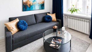 A modern living room with a grey sofa, blue and beige cushions, a glass coffee table with magazines, and a cityscape painting on the wall. - Elegant Apartment near Vitsula River (Warszawa)