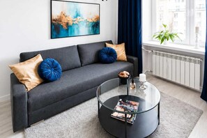 A modern living room with a grey sofa, blue and beige cushions, a glass coffee table with magazines, and a cityscape painting on the wall. - Elegant Apartment near Vitsula River (Warszawa)