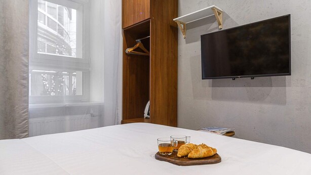 Une chambre d’hôtel avec un lit, un téléviseur fixé au mur et un plateau de petit-déjeuner comprenant des croissants et du thé.