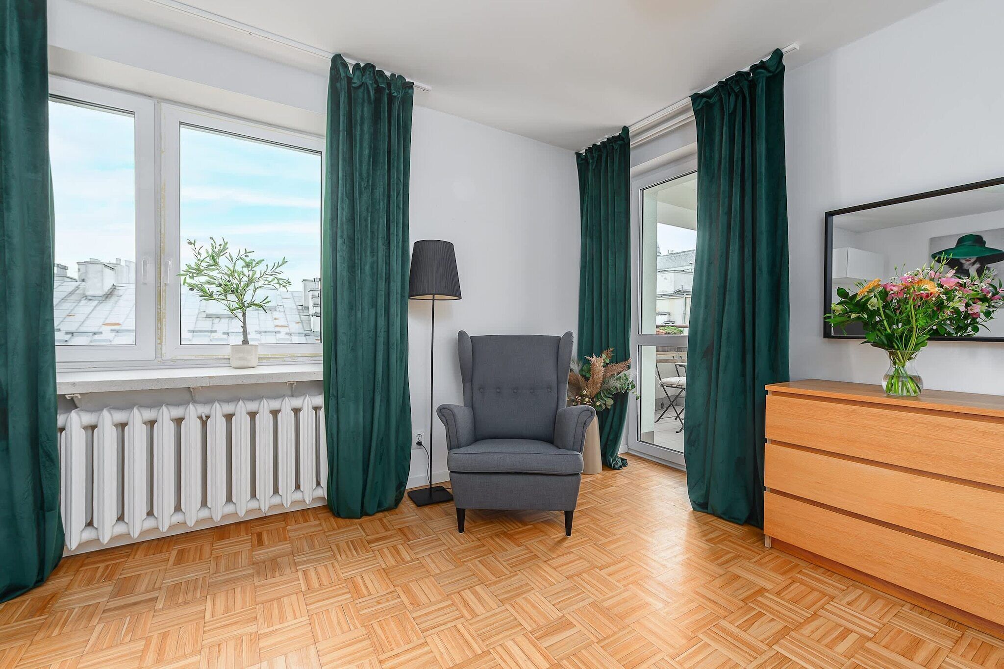 Une pièce avec un radiateur, un fauteuil gris, une commode en bois et un lampadaire.