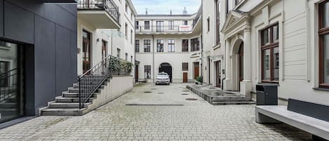 A courtyard with a paved walkway, a car parked in the center, and buildings with balconies and windows.