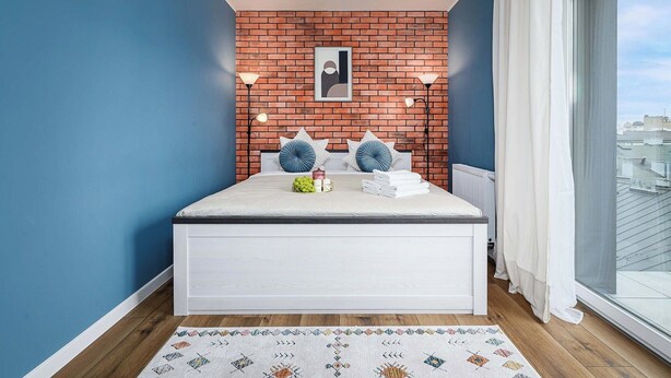 A bedroom with a white bed, a brick accent wall, a large window, and a view of buildings. - Home near Piotrkowska Street (Łódź)