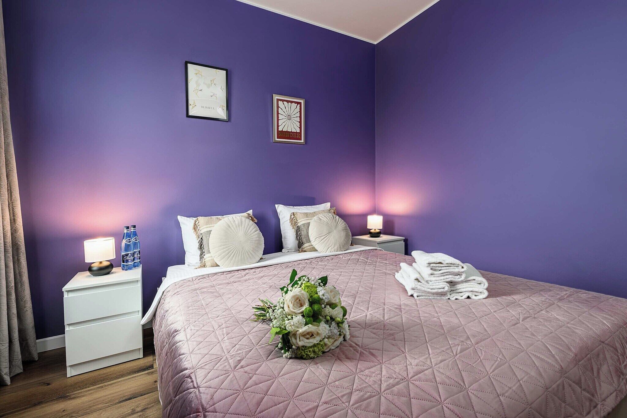 Una camera da letto con un letto, cuscini, un comodino con una lampada, una parete con due quadri e un bouquet di fiori.
