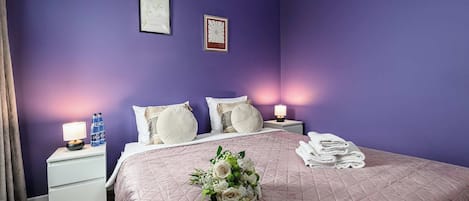 Un dormitorio con cama, almohadas, una mesita de noche con lámpara, una pared con dos cuadros enmarcados y un ramo de flores.