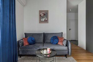 Een woonkamer met een grijze bank, blauwe en rode kussens, een klein rond salontafeltje met een dienblad, en een lijstwerk aan de muur.