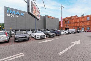 Ein Parkplatz mit mehreren Autos, ein Kinogebäude mit 'IMAX'-Beschilderung und ein rot backsteinsches Gebäude mit der Aufschrift 'CINEMA CITY'.