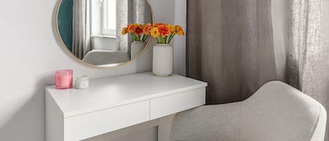 A white vanity with a round mirror, a vase with flowers, and a pink candle.