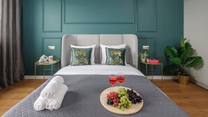 A neatly made bed with a grey quilt, two pillows with a floral design, a bedside table with a lamp, a potted plant, and a tray with grapes and wine glasses. - Magnificent seafront duplex in Porspoder (Łódź)