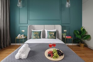 Ein ordentlich bezogenes Bett mit einer grauen Tagesdecke, zwei Kissen mit Blumenmuster, ein Nachttisch mit Lampe, eine Topfpflanze und ein Tablett mit Trauben und Weingläsern.