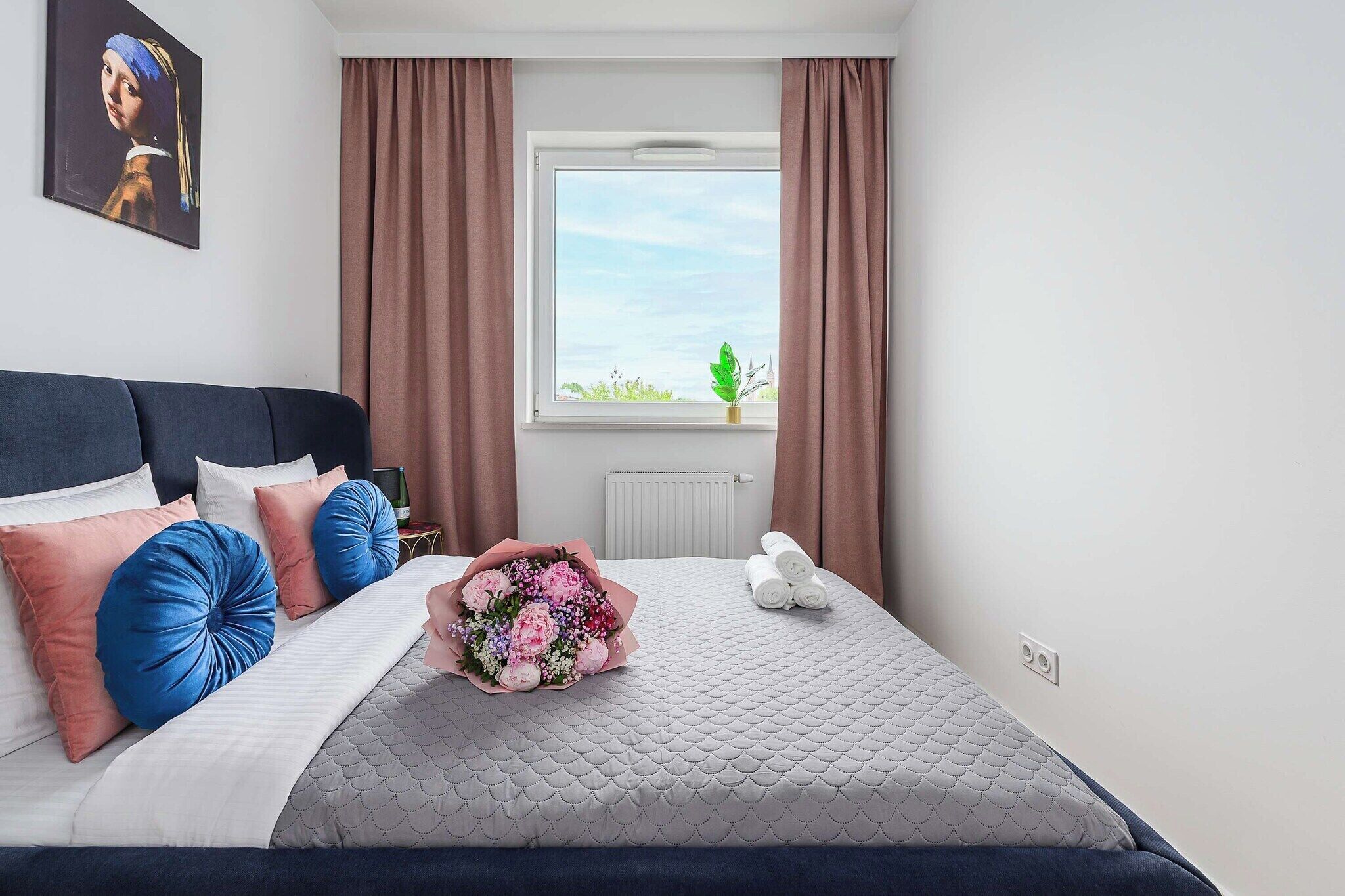 A bedroom with a bed, pillows, a bouquet of flowers, a radiator, and a window with curtains.