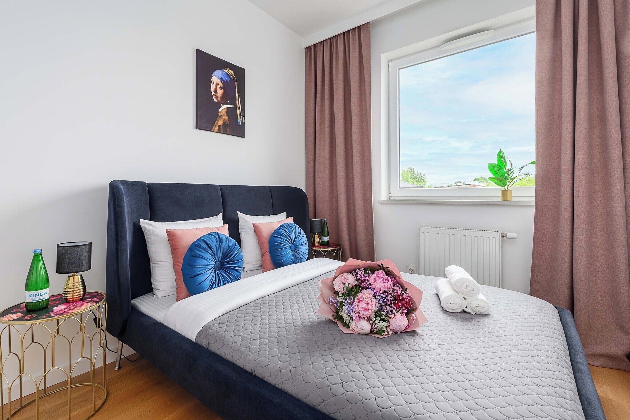 Ein modernes Schlafzimmer mit einem großen Bett, einem Fenster mit Vorhängen, einem Heizkörper und einem kleinen Tisch mit Lampe und Flasche.