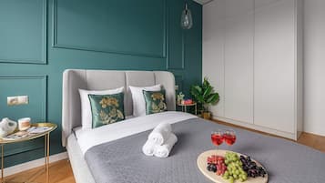 A neatly made bed with a grey quilt, two pillows with floral embroidery, and a tray with grapes and wine glasses on the bedside table.