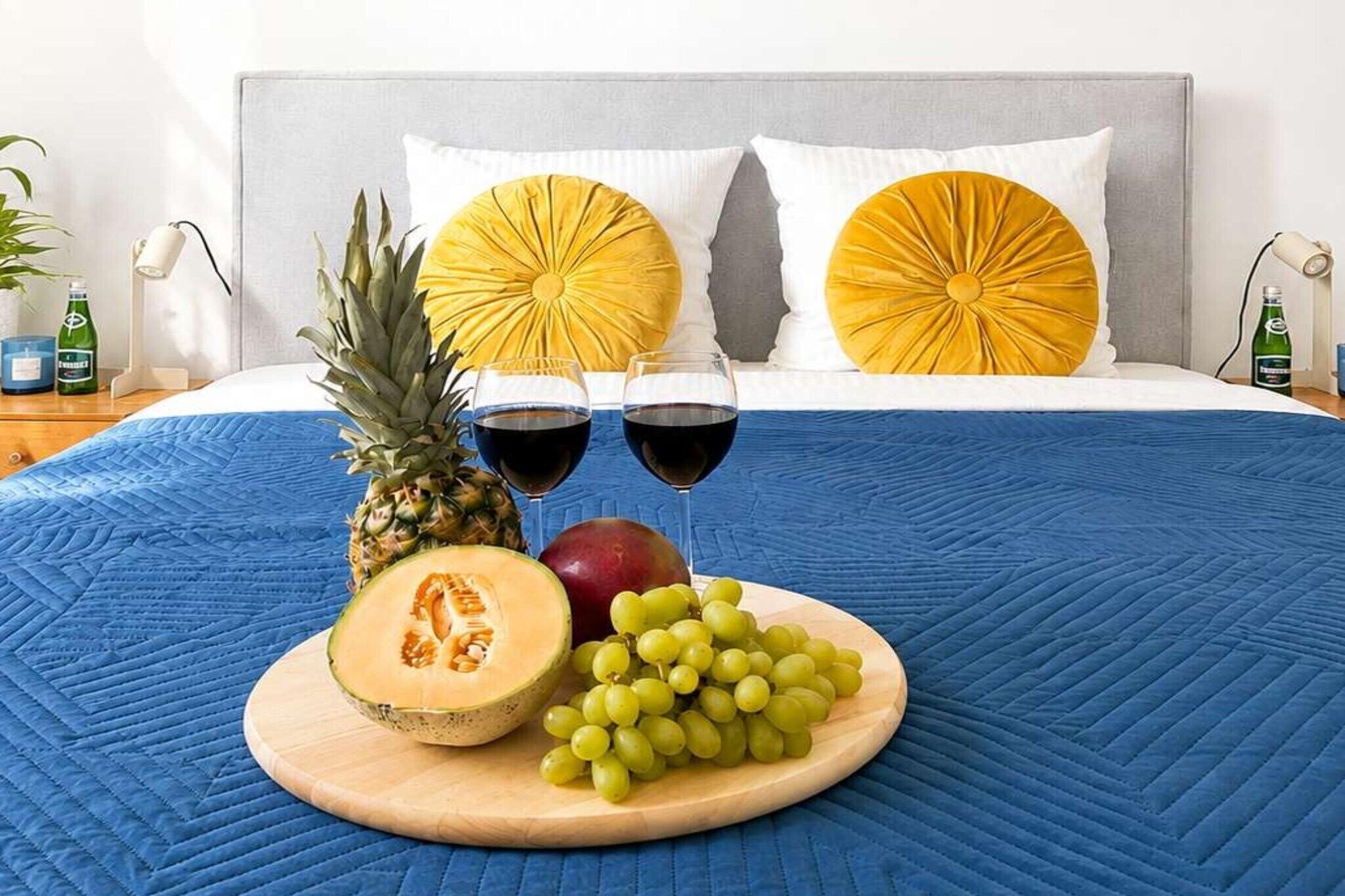 Ein Bett mit einer blauen Steude, zwei Gläser Rotwein, eine Ananas, eine halbierte Melone, ein roter Apfel und eine Schale mit grünen Trauben.