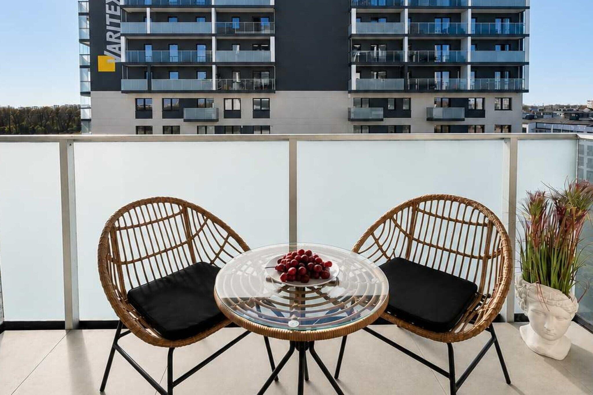 Un balcon avec deux fauteuils en osier et une table en verre, une plante en pot et un bâtiment en arrière-plan.