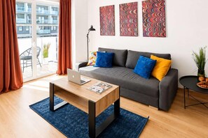 Ein modernes Wohnzimmer mit einem grauen Sofa, blauen und gelben Kissen, einem Holztisch mit einem Laptop und einem blauen Teppich.