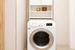 A front-loading washing machine with a control panel, a shelf above it, and a wooden door to the left. - 3-Room Home near Atlas Arena (Łódź)