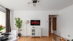 Uma sala de estar moderna com uma TV de tela plana exibindo 'NETFLIX', uma área de jantar com mesa posta para dois, uma planta em vaso e um abajur.