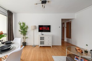 Ein modernes Wohnzimmer mit einem Flachbildfernseher, der 'NETFLIX' anzeigt, einem Essbereich mit Tisch für zwei Personen, einer Topfpflanze und einer Stehlampe.