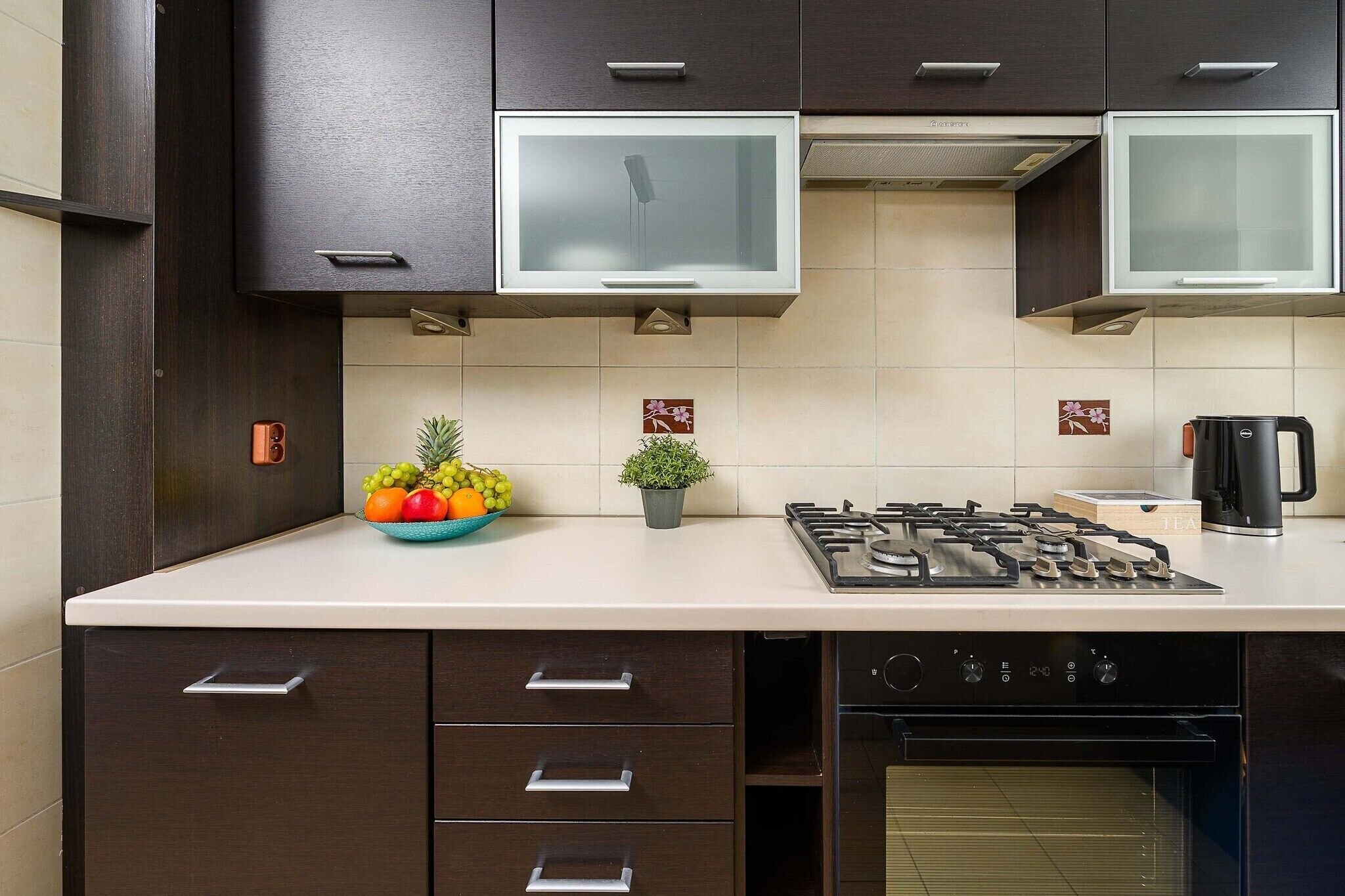 A kitchen with dark cabinets, a gas stove, an oven, and a bowl of fruit on the counter.