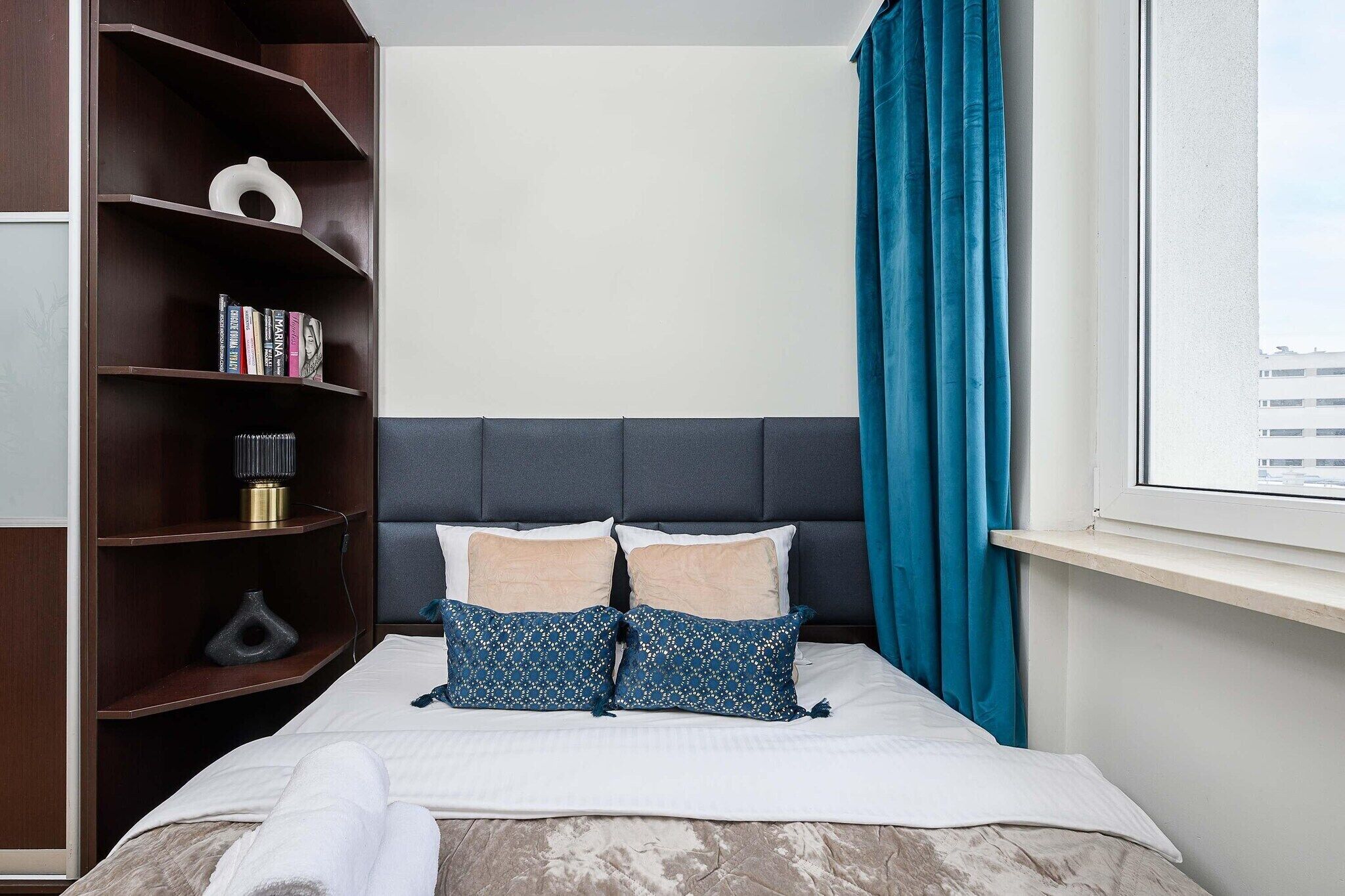 Una cama bien hecha con dos almohadas, una mesita de noche, una estantería y una ventana con cortinas.