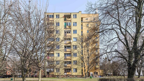 Um edifício residencial com vários andares e varandas, rodeado por árvores sem folhas e um campo desportivo cercado.