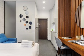 A hotel room with a bed, a desk, a chair, a wooden panel wall, a mirror, and a clock on the wall. - Stylish City Apartment for 3 Guests (Warszawa)