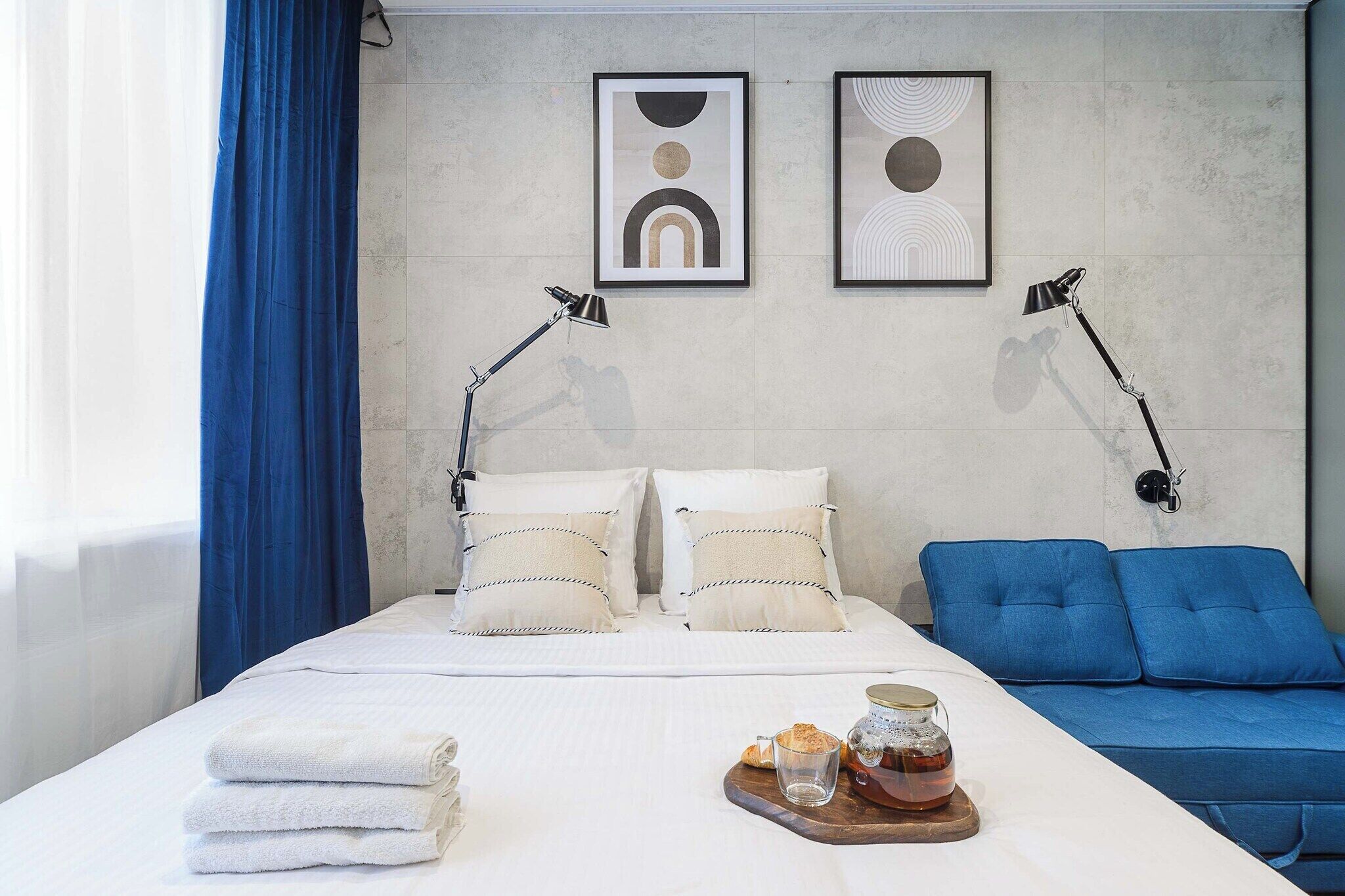 A hotel room with a bed, two bedside lamps, a blue sofa, a tray with bread and a drink, and two abstract wall art pieces.