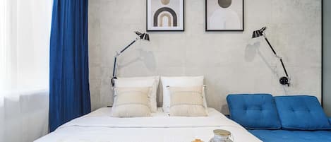 A hotel room with a bed, two bedside lamps, a blue sofa, a tray with bread and a drink, and two abstract wall art pieces.