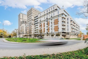 A multi-story residential building with balconies, a landscaped area with trees, and a road in the foreground. - Cozy Flat near Manufaktura (Łódź)