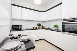 A modern kitchen with white cabinets, a black countertop, a microwave, an oven, and a dining area set with plates, glasses, and cutlery. - Cozy Flat near Manufaktura (Łódź)