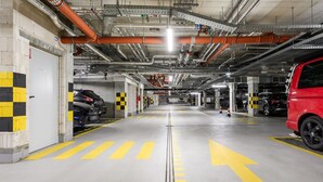 Un estacionamiento subterráneo de varios niveles con flechas direccionales claras en el suelo, múltiples espacios de estacionamiento y un vehículo rojo estacionado a la derecha.