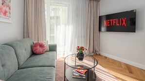 Una sala de estar con un sofá verde, una mesa de centro de vidrio, un televisor de pantalla plana y una ventana con cortinas translúcidas.