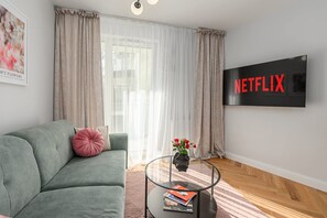 Ein Wohnzimmer mit einem grünen Sofa, einem Glastisch, einem Flachbildfernseher und einem Fenster mit transparenten Vorhängen.