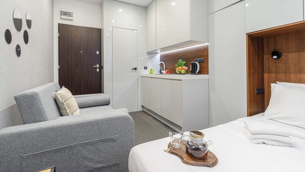 A modern hotel room with a grey sofa, a bed with white linens, a small kitchen area, and a wooden headboard. - Stylish Urban Apartment for 3 (Warszawa)