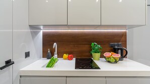 A modern kitchen with a white countertop, a built-in sink, a faucet, a black induction cooktop, a coffee maker, a potted plant, and fresh vegetables. - Stylish Urban Apartment for 3 (Warszawa)