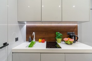 A modern kitchen with a white countertop, a built-in sink, a faucet, a black induction cooktop, a coffee maker, a potted plant, and fresh vegetables. - Stylish Urban Apartment for 3 (Warszawa)