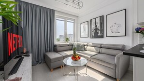 Una sala moderna con un sofá seccional gris, una mesa de centro de vidrio con fruta y un ventanal con cortinas.