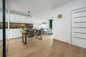 A modern kitchen with white cabinets, a dining table with chairs, and a living area with a sofa and a green curtain. - Charming House in Landéda with Sea View (Łódź)