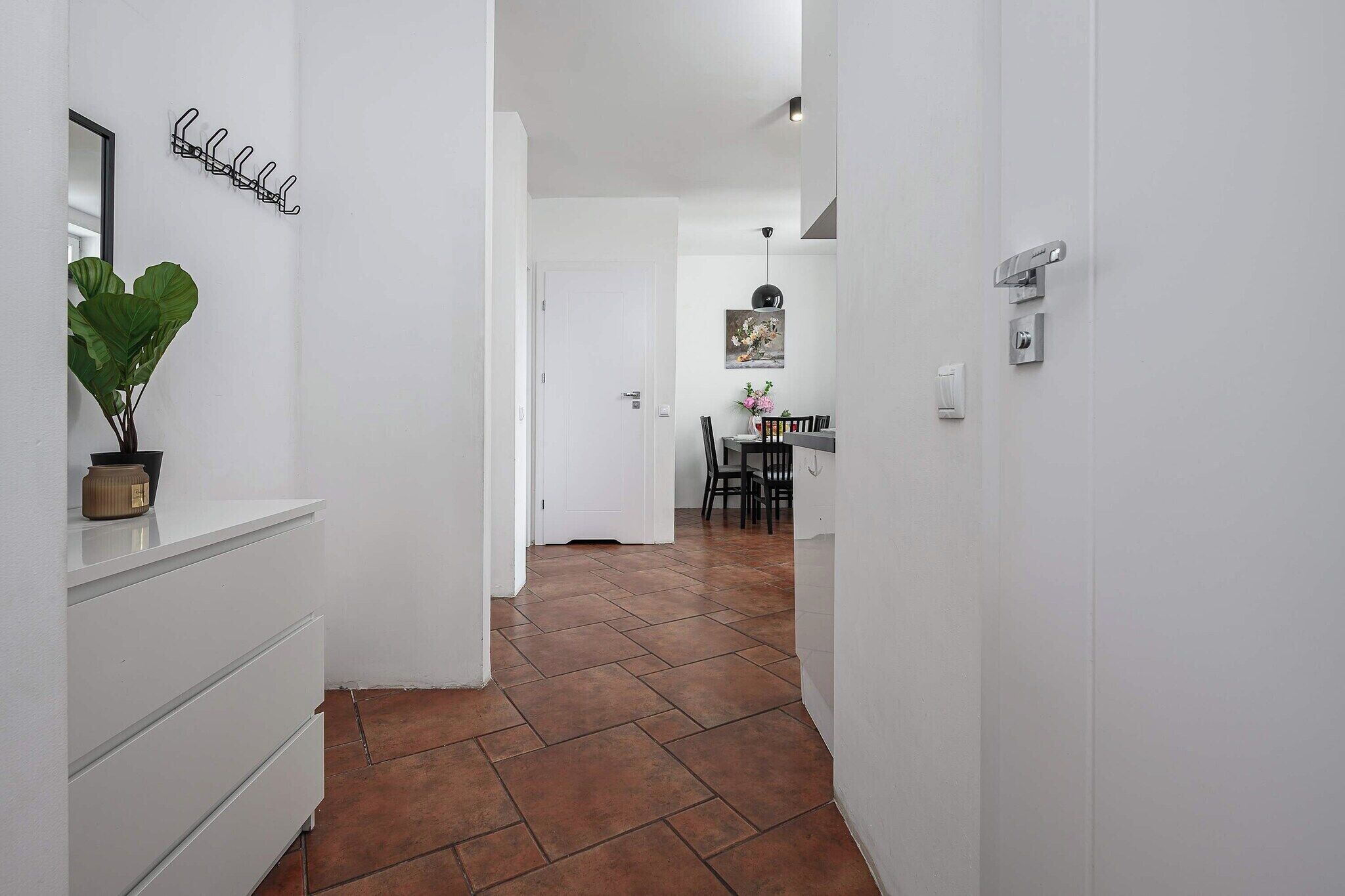 A hallway with a tiled floor, a potted plant, a coat rack, and a dining area in the distance.