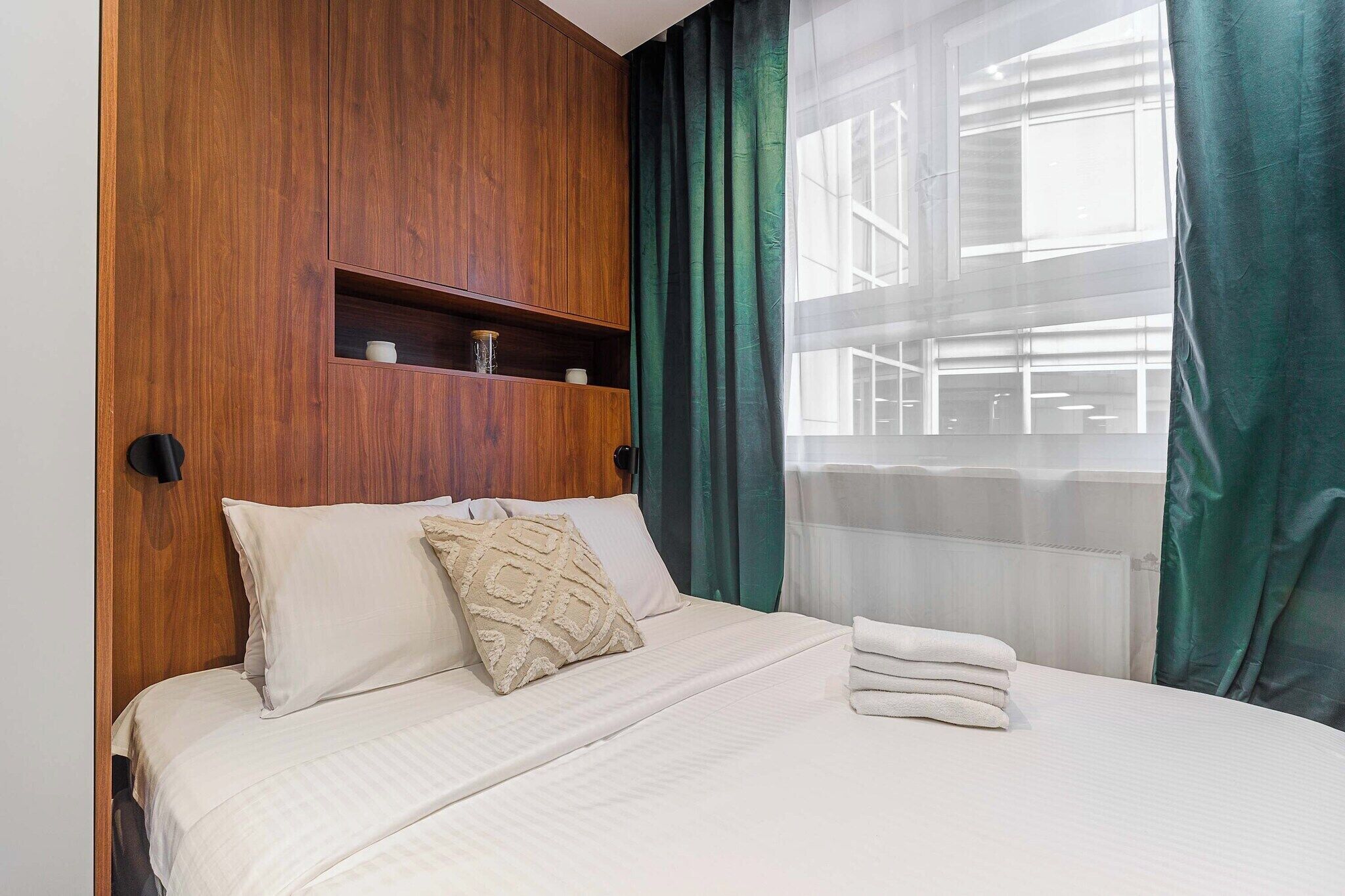 Une chambre d’hôtel avec un lit, une tête de lit en bois, une étagère avec des objets décoratifs et une vue à travers des rideaux transparents.